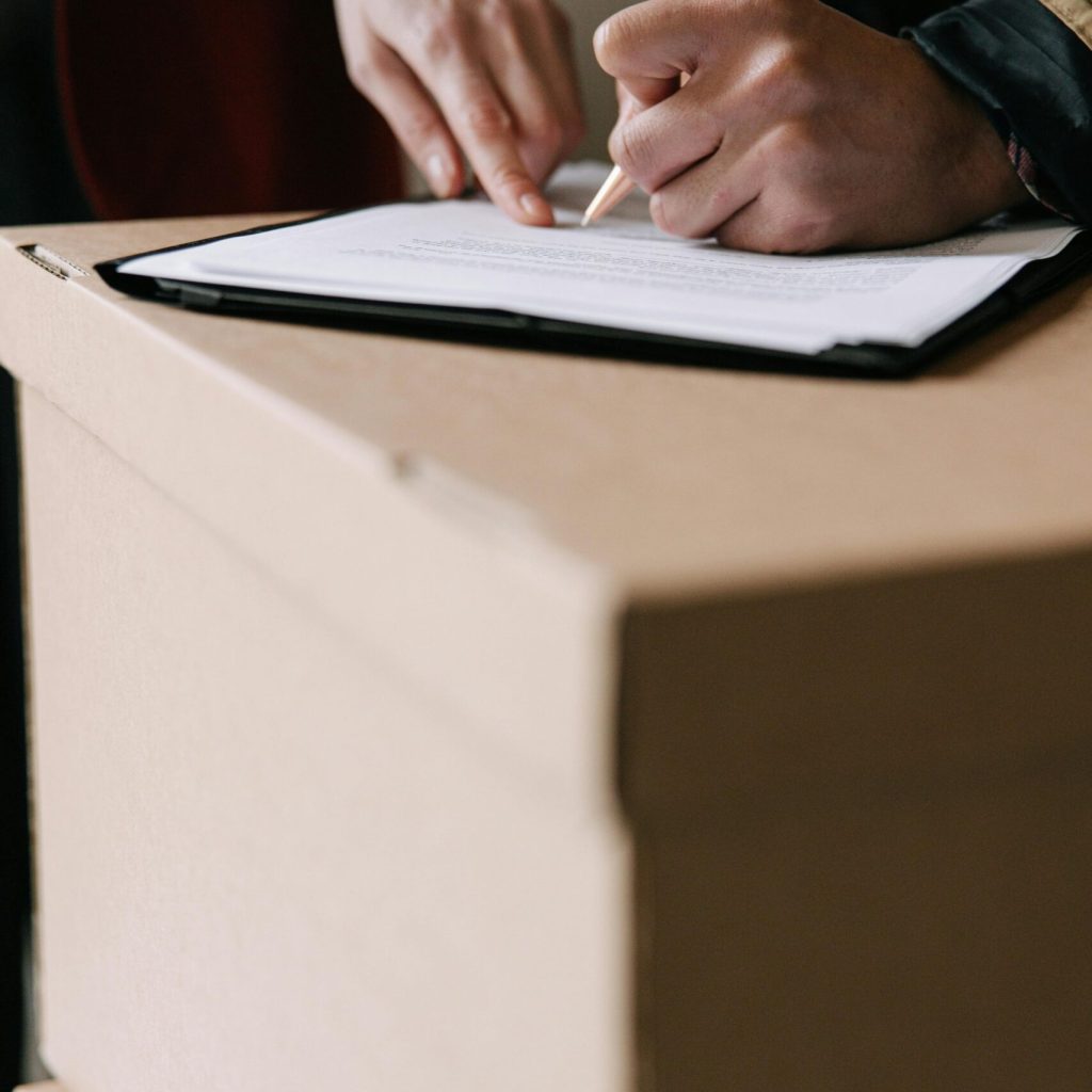 pexels-photo-8962685-8962685 Hands signing lease documents on a cardboard box in an apartment.
